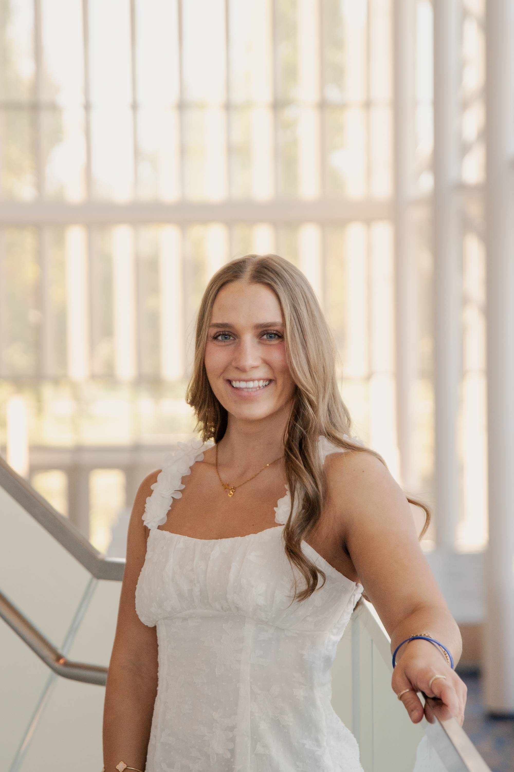 woman with long blonde hair wearing a white dress smiling at the camera with tall windows in the background